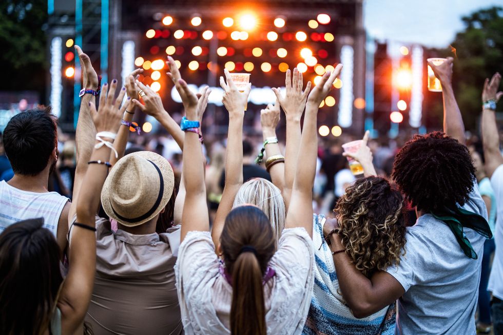 Audience enjoying an outdoor music festival, raising hands and drinks towards the stage with bright lights.