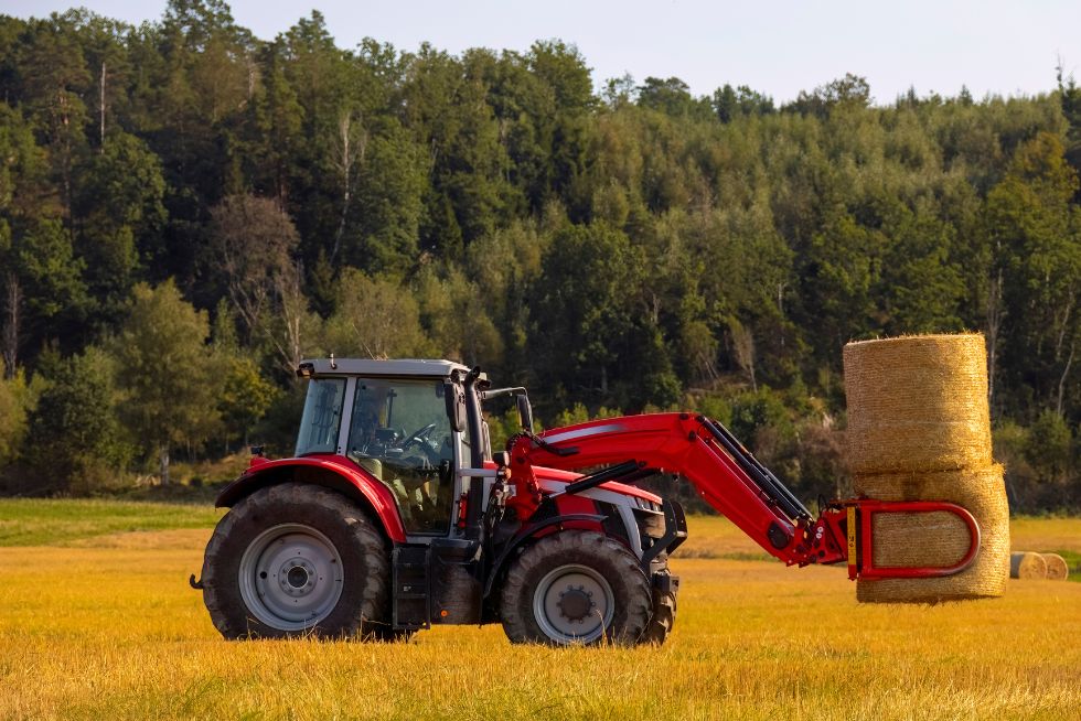 Red tractor lifting hay bales in a sunny field with a forest background.