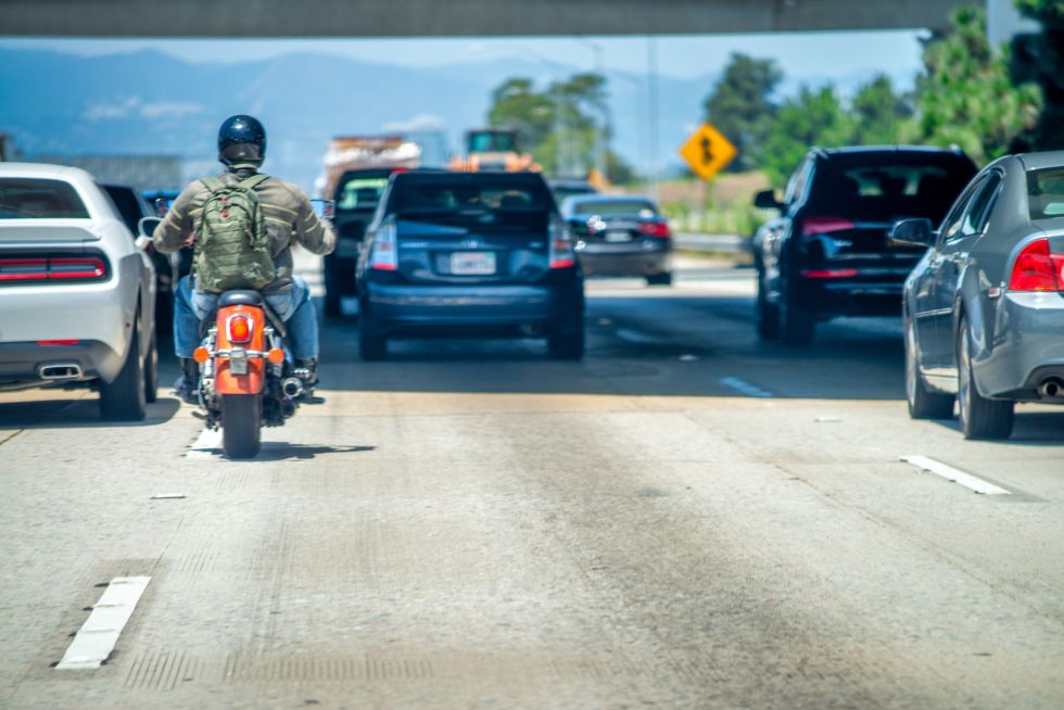 Motorcyclist rides between cars on a busy highway under clear skies, showcasing lane-splitting in traffic.