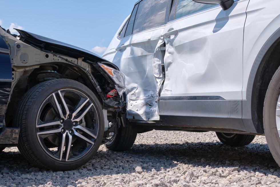 Two cars involved in a side-impact collision on a gravel road under a clear blue sky.