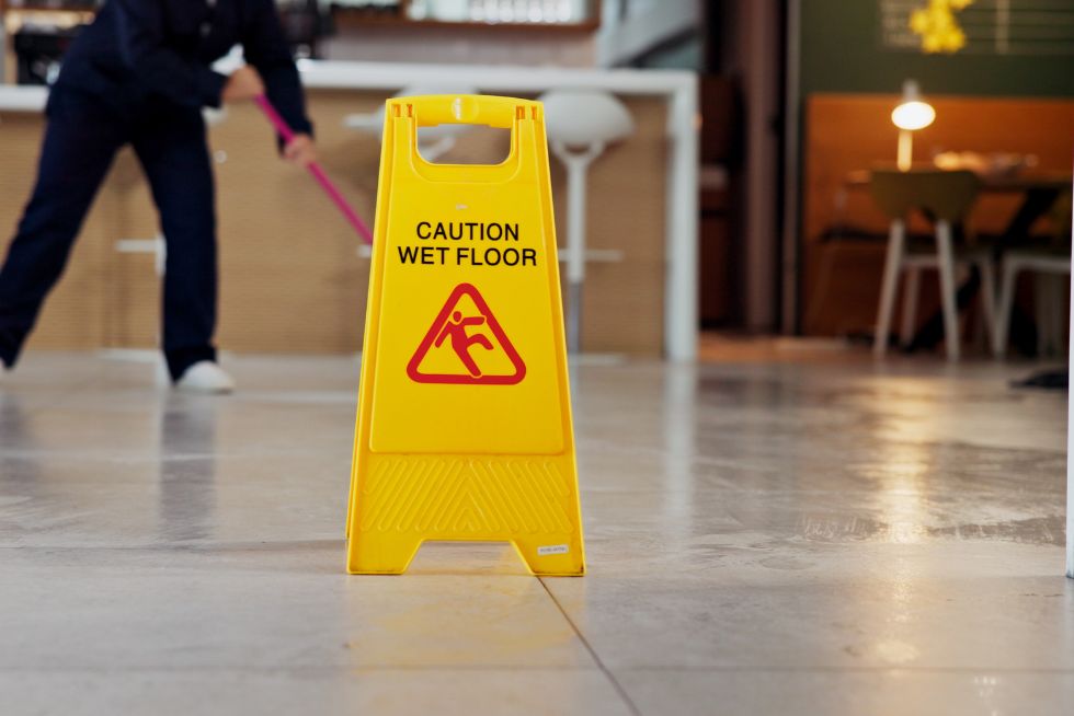 Yellow sign warns Caution Wet Floor while a person mops in the background, ensuring safety at the workplace.