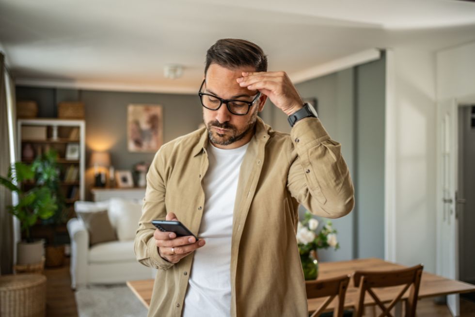 Man in glasses looks at smartphone, appearing thoughtful in a modern living room setting.
