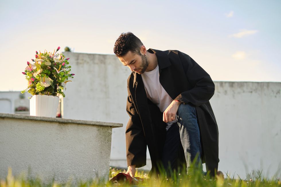 Man kneeling at grave with flowers, wearing black coat, sunny day.